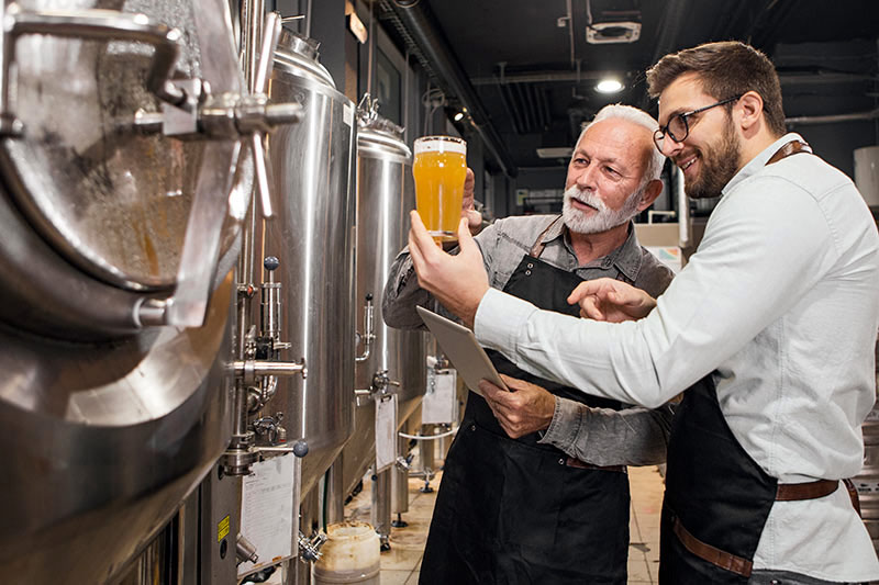 two brewery workers examining glass of beer next to large stainless steel beer kettles
